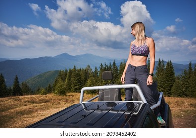 Chica atlética con sostén deportivo de colores parada en el cuerpo de un SUV negro frente a los asientos de los pasajeros y mirando a la izquierda en el fondo del bosque verde, colinas montañosas y hermoso cielo nublado de verano.