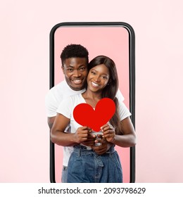 Amor y cuidado. Retrato de una feliz pareja negra abrazándose sosteniendo un corazón de tarjeta de papel rojo parado dentro de la pantalla del teléfono celular aislado en el fondo del estudio rosa, un tipo sonriente abrazando a una mujer. Día de San Valentín