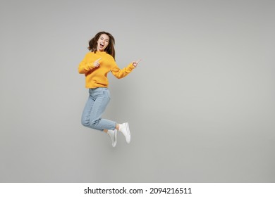 Toda la longitud de una joven emocionada y expresiva mujer con suéter amarillo de punto corre rápido en el aire salta con el dedo índice a un lado en el espacio de trabajo simulado aislado en un retrato de estudio de fondo gris.