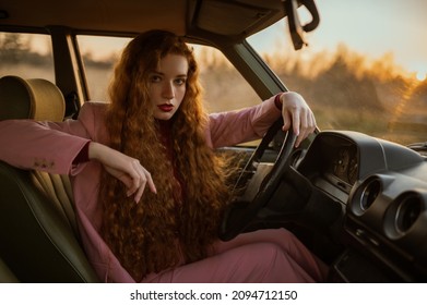 Mujer segura de moda posando al volante de un coche retro verde. Elegante modelo pelirroja con traje rosa clásico. Copiar, espacio vacío para texto