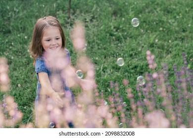 Partially blurred little girl 3-4 with dark hair in denim dress catches soap bubbles on the lawn near flowers