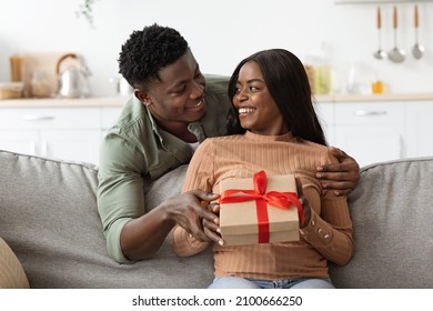 Liefdevolle Afro-Amerikaanse man die zijn vrouw knuffelt, een geschenkdoos geeft, een grote verrassing maakt voor St. Valentijnsdag, interieur, vrije ruimte. Gelukkige zwarte man die zijn vriendin omhelst en cadeau vasthoudt