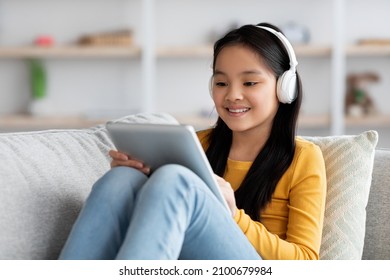 Zrelaksowane chińskie dziecko preteen ładna dziewczyna odpoczywa na kanapie z cyfrowym tabletem, nosi bezprzewodowy zestaw słuchawkowy, ogląda film animowany lub miły dla dzieci w Internecie, wnętrze domu, kopia przestrzeń