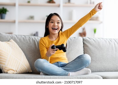 Ragazza asiatica carina e felice, studentessa seduta sul divano in soggiorno, che tiene in mano un joystick, gioca a un videogioco portatile e gesticola, si gode il fine settimana a casa, concetto di intrattenimento per bambini, spazio di copia