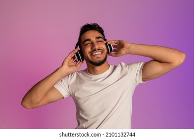 Peaceful young Arab guy listening to music in wireless headset, closing eyes and relaxing in neon light. Portrait of positive middle Eastern man enjoying favorite playlist or audio book