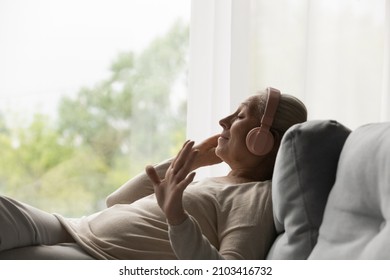 Side view happy old mature retired woman listening popular music in headphones, relaxing alone on cozy sofa, enjoying peaceful carefree weekend time alone at home, stress free leisure pastime.