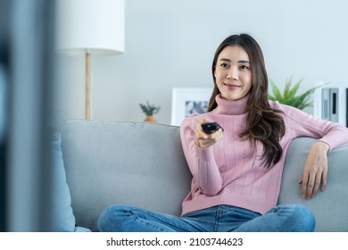 Wanita muda Asia yang cantik sedang menonton film seru di televisi di rumah. Gadis kasual yang menarik merasa senang dan rileks, duduk di sofa sambil bersenang-senang menonton video komedi di TV di rumah. Konsep gaya hidup yang aktif