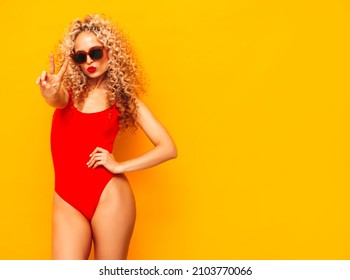 Young beautiful smiling woman posing near blue wall in studio.Sexy model in red swimwear bathing suit.Positive female with afro curls hairstyle.Shows peace sign.Happy and cheerful