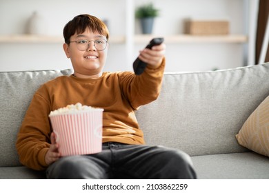 リラックスしたかわいい太りすぎの中国の子供の 10 代の少年はポップコーンでいっぱいのバスケットでソファに座って、リモコンを持って、家でテレビを見て、週末の自由な時間を楽しんで、スペースをコピー