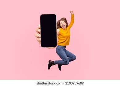 Happy young woman flying and jumping in air and showing big mobile empty screen for copy space and advertising area. indoor studio shot isolated on pink background
