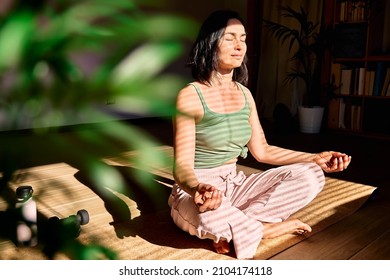 Frau, die zu Hause Yoga und Meditation praktiziert und in Lotus-Pose auf einer Yogamatte sitzt, entspannt mit geschlossenen Augen. Achtsames Meditationskonzept. Wohlbefinden.