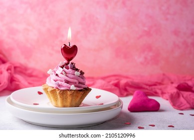 Cupcake mit rosa Creme und roter herzförmiger Kerze auf rosa Hintergrund, Essen zum Valentinstag.