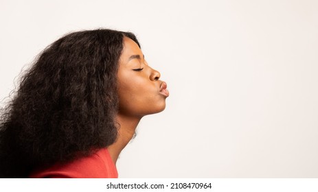 Imagen de perfil de una joven negra haciendo pucheros con los labios sobre un fondo blanco de estudio, una mujer afroamericana milenaria romántica parada con los ojos cerrados, lista para besarse, vista lateral, panorama con espacio para copiar