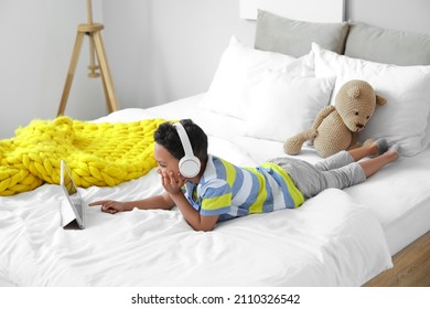 Little African-American boy with headphones watching cartoons on tablet computer in bedroom