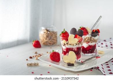 Sweet breakfast on Valentine's Day on a white wooden table. Creamy dessert with ripe strawberries and pureed raspberries, muesli in a glass decorated with chocolate in the shape of a heart. Side view