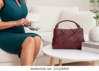 Một phụ nữ trẻ xinh đẹp tạo dáng trong chiếc váy màu xanh lá cây trên nền ghế sofa trong phòng khách. Một người phụ nữ đang cầm một chiếc túi đeo vai màu đỏ tía làm bằng da bện. Khái niệm về thời trang của phụ nữ.