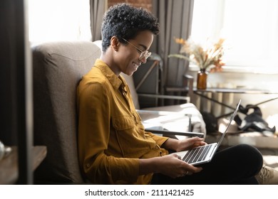 Wanita Afrika milenium berambut pendek yang bahagia menggunakan komputer laptop, menonton film, berbicara di panggilan video, tertawa, menikmati hiburan digital, waktu luang, duduk di sofa di rumah