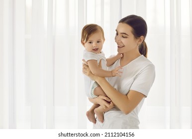 Portret in lichte kamer van jonge liefhebbende moeder die haar mooie zes maanden oude dochter vasthoudt. Moeder staat op de achtergrond van een groot helder raam met witte tule die teder naar de baby kijkt. Ruimte kopiëren.