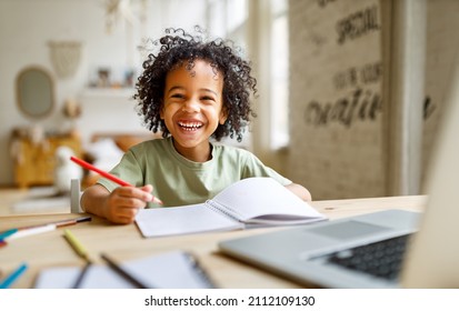 Formazione a distanza. Un bambino afroamericano sorridente studia online sul portatile a casa, seduto al tavolo e comunica con l'insegnante tramite videochiamata sul computer.