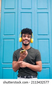 Millennial hipster male with afro hair, beard and earring, enjoys a music podcast with yellow headphones outdoors. Biracial curly haired man smiles looking at the camera on blue background, copy space