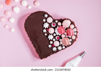 Draufsicht auf rosa herzförmigen Kuchen mit Schokoladenglasur, Meringues und Makronen als Dekoration. Auf rosa Hintergrund platziert. Valentinstag, Muttertag, Geburtstag, Feierkonzept.