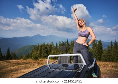 Mujer joven en sujetador deportivo brillante con expresión de cara feliz de pie en el cuerpo del camión y sosteniendo una mano en la cintura y otra levantando. Hermosos paisajes de montaña en el fondo.