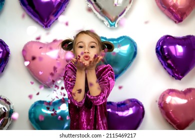Mädchen bläst rosa Konfetti in Form von Herzen aus ihren Handflächen, während sie in die Kamera schaut. Feiern Sie am Valentinstag mit Folienballon-Dekorationen. Bunte brillante, glänzende Ballons