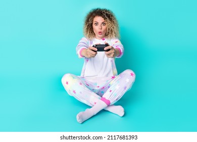 Ganzkörperfoto einer beeindruckten, tausendjährigen Dame mit welliger Frisur, die eine Playstation trägt und Pyjamasocken auf blauem Hintergrund isoliert trägt