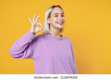 Smiling fun young blonde caucasian woman with bob haircut showing okay ok gesture wearing basic purple shirt looking camera charming toothy smile isolated on yellow color background studio portrait