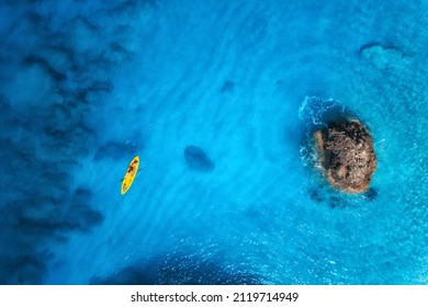 Pemandangan udara kayak kuning di laut biru saat matahari terbenam di musim panas. Pria di kano mengambang di air biru jernih, batu, batu. Pulau Lefkada, Yunani. lanskap tropis. papan sup. Perjalanan aktif. Pandangan atas