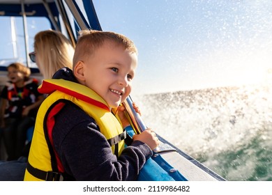 Porträt des kleinen blonden, glücklichen, aufgeregten, lächelnden kaukasischen Jungen, der eine Schwimmweste trägt, genießen Sie das Segeln auf dem Motorboot-Meer gegen den blauen Himmel und die Wasserspritzer-Wellensonne mit Hintergrundbeleuchtung. Erholung im Sommerurlaub