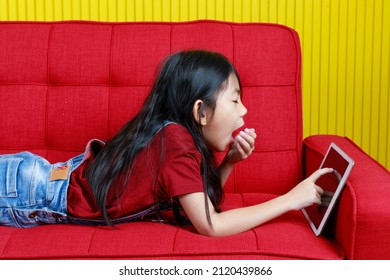 デニム ジーンズのオーバー オールの衣装でアジアの眠そうな若い小学生の女の子のクローズ アップ ショットは、タッチ スクリーン タブレット コンピューターでサーフィン インターネットを閲覧しながら手カバー口あくびを保持している横になっています。