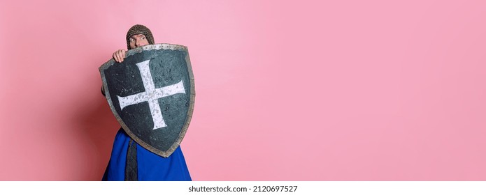 Retrato de um homem assustador, guerreiro medieval ou cavaleiro em armadura de malha, escondendo-se atrás de um escudo isolado sobre um fundo rosa de estúdio. Comparação de épocas, história e estilo renascentista.