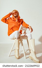 Mulher estilosa usando um elegante moletom laranja com capuz, óculos de sol, calça jeans skinny branca e botas de cano alto, sentada e posando em uma cadeira. Retrato de corpo inteiro em estúdio.