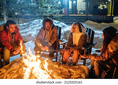 Groep gelukkige vrienden die op stoelen zitten en marshmallows koken bij het vuur tijdens hun wintervakantie in het landhuis