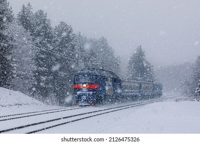 A diesel locomotive with an intercity passenger train rushes through a snow storm along a railway line through a forest. Winter snowy weather. Railway passenger transportation.