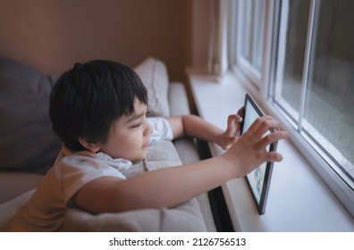 Filmisch portret van een gelukkige jonge jongen die online een spel speelt op de tablet, Kid kijkt naar tekenfilm van internet, Kind zit op de bank naast het raam met een regenachtige dag in de ochtend