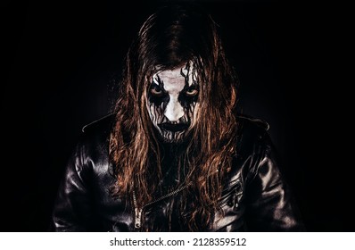 長い茶色の髪と塗られた顔を持つ黒い金属のメタルヘッドの男のポートレート写真は、革のジャケットに立って、黒い背景にカメラを見ています。