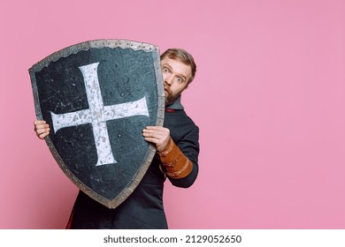 Retrato de um homem assustador, guerreiro medieval ou arqueiro em armadura, escondendo-se atrás de um escudo isolado sobre um fundo rosa de estúdio. Lutador assustado. Comparação de eras, história e estilo renascentista.