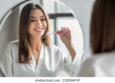 Hermosa mujer joven cepillando los dientes mientras está de pie cerca del espejo en el baño, feliz mujer milenaria sosteniendo cepillo de dientes y sonriendo a su reflejo, haciendo higiene matutina en casa, enfoque selectivo