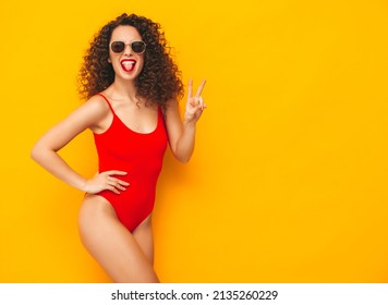 Young beautiful smiling woman posing near yellow wall in studio.Sexy model in red swimwear bathing suit.Positive female with curls hairstyle.Happy and cheerful. In sunglasses. Showing peace sign