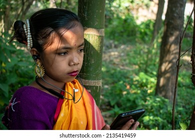 A tribal little girl is listening to music alone with Bluetooth headphones on her android phone in the forest. She is wearing traditional aboriginal attire.