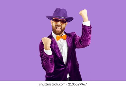 成功を祝う幸せな自信のある男。帽子、スーツ、紫色の背景に立っているオレンジ色の蝶ネクタイを着た面白い興奮した陽気なひげを生やした男は、拳を上げて叫びます