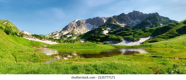 Tienda de campaña en la verde orilla floreciente de un lago de montaña rodeado de picos de montaña. Adygea, LagoNaki, lago psenodakh