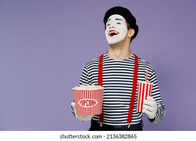 Lachender, erstaunlicher, wunderbar lebhafter junger Mime-Mann mit weißer Gesichtsmaske trägt gestreiftes Hemd, Baskenmütze, alten Eimer Popcorn, Tasse Limonade, isoliert auf schlichtem pastellfarbenem hellviolettem Hintergrund, Studioporträt