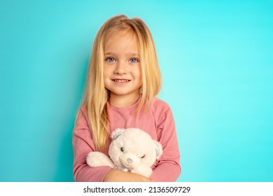 En lille sød pige med store blå øjne og blond hår holder en bamse i hænderne. Et barn i en lyserød sweater på en blå baggrund med et yndlingslegetøj.