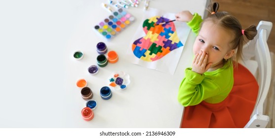 女の子は自閉症の日のはがきを描き、世界中のすべての子供たちにエアキスを送ります. チューブに入ったガッシュと水彩絵の具と、テーブルの上のパレット。クリエイティブな赤ちゃんが描いたパズルハート