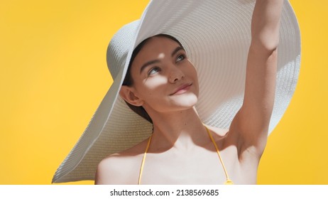 Una chica de cabello castaño con un gran sombrero blanco se cubre la cara del sol con la palma de la mano mirando hacia el cielo sobre un fondo amarillo. Anuncio de protección de la piel y cuidado del cuerpo.