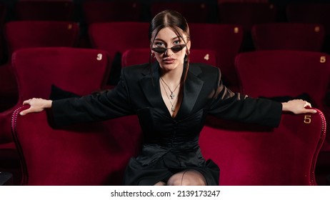 Porträt eines ganz in Schwarz gekleideten Models, das auf einem roten Kinostuhl mit schöner Sonnenbrille sitzt und in die Kamera blickt