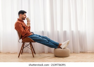 Rest Concept. Happy Arab guy drinking coffee sitting on chair in living room. Casual man relaxing, enjoying weekend free time or break from work, free copy space, profile side view, full body length